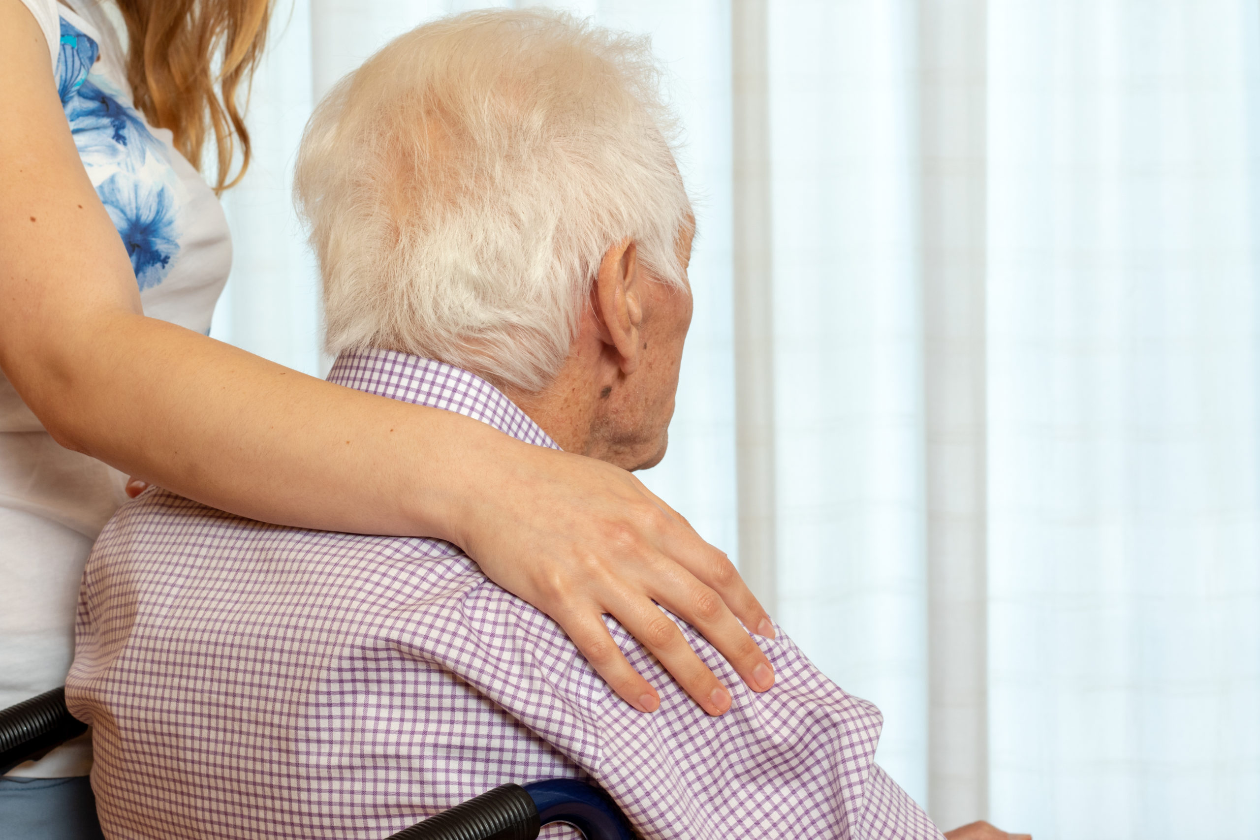 Close up rear view detail of female assistant with hand on old man’s shoulder. Senior man in wheelchair looking at window.
